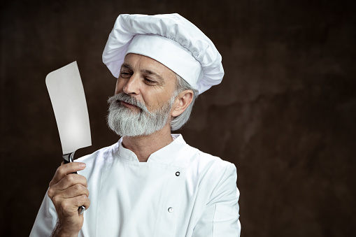 Adult man in a chef's whites, white hat and white double-breasted jacket looking at a knife. Advertisement portrait for butchery or knives.