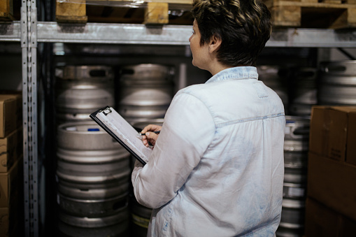 Mid adult woman manager with clipboard in brewery distribution warehouse checking supplies
