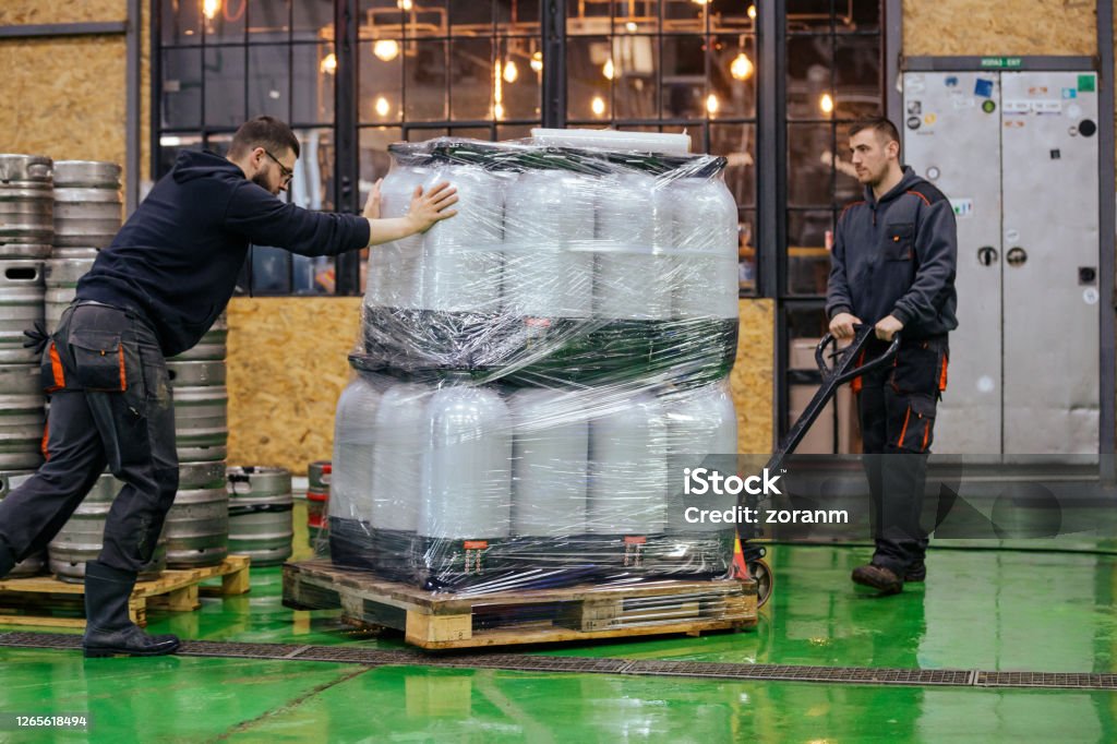 Two men moving pallet jack loaded with kegs in brewery warehouse Two young man working in brewery warehouse, pushing pallet jack loaded with kegs wrapped in plastic foil Beer - Alcohol Stock Photo