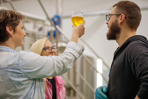 Woman in brewery production line, holding up a glass of fresh craft beer and examining it with colleagues