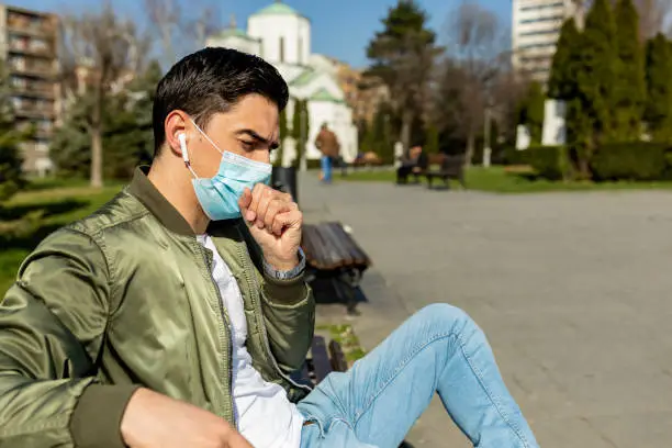 Photo of COVID-19 Man in city street wearing face mask protective for spreading of Coronavirus Disease in europe. Portrait of man with surgical mask on face against SARS-CoV-2. Tired man with a mask on his face sits on a bench listening to music through headphones