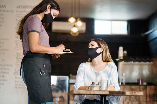 A latin waitress wearing a face mask and taking an order of a female client with mask.