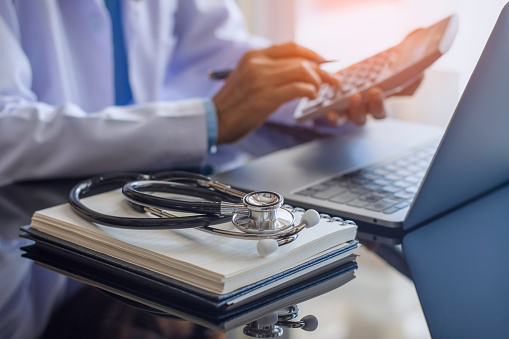 Female doctor or practitioner using calculator and work on laptop computer with medical stethoscope  and notebookon the desk at clinic or hospital. Medical healthcare costs ,fees and revenue concept.