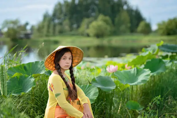 Beautiful teen girl looking at the camera. Beautiful girl with Ao-Dai traditional Vietnam dress costume for woman. Beautiful nature. A girl with two pigtails in a straw hat sits on the shore of the lake next to a blossoming lotus flower