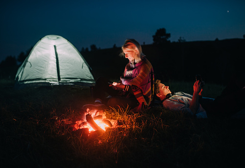 Young couple working around the campfire on  their smartphones near tent and
they warm themselves by the fire at night in the mountains.
Technology in nature.