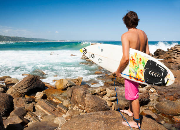 Garden Route, South Africa - March 2, 2013: A male surfer holds his surfboard while  standing on rocks looking at waves, Victoria Bay Beach, Western Cape, Africa