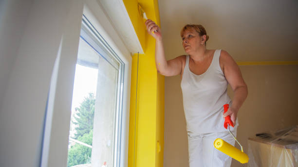 Woman in protective suit painting wall and ceiling with paint roller in black color during interior renovation