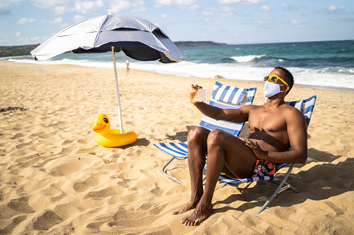 Lonely afro man with protective face mask sitting on a lounge chair on an empty beach and making selfie