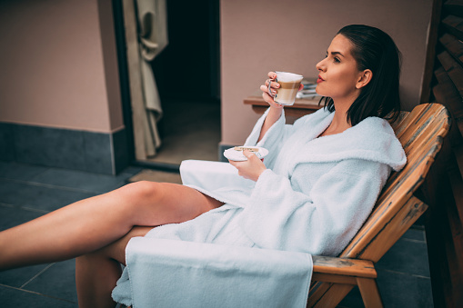 Beautiful young woman wearing a bathrobe sitting on the balcony, drinking and enjoying morning cup of coffee