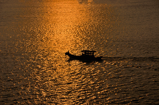 A fisherman sits in silhouette on the bow of his family's sampan in the Mekong Delta at sunset with the sun turning the water into gold