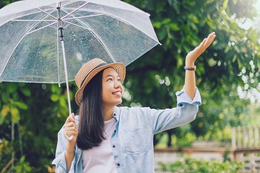 Asian beautiful smiling woman covering umbrellas in the rain with hand playing rain drop in the park with green tree background.Concept of preparing for the rainy season.