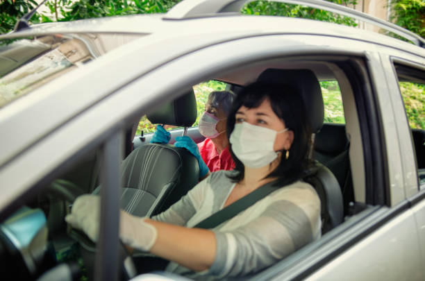 Caregiver drives a senior adult woman in a white car for daily medical procedures. Both put on protective masks. stock photo
