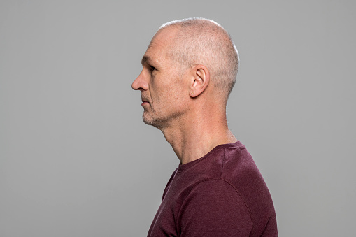 Profile of mature man in maroon t-shirt standing against grey background.