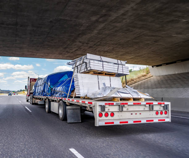 istock Red big rig semi truck transporting covered and tightened cargo on flat bed semi trailer running under the bridge across wide highway 1265921197
