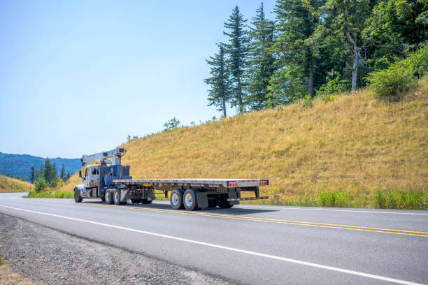 istock Big rig semi truck with crane application on the frame transporting empty flat bed semi trailer running on the winding road with yellow summer hills 1265921295