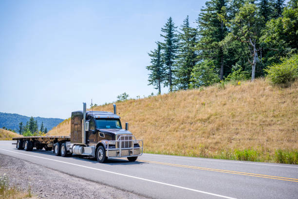 istock Powerful big rig black semi truck with grille guard going up hill with empty flat bed semi trailer in gorgeous Columbia Gorge area 1265922014