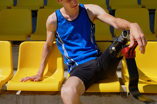 Close up portrait of adaptive athlete with prosthetic foot sitting on the empty stadium on sunny summer day. Real bodies, equal sport, disability recovery concept
