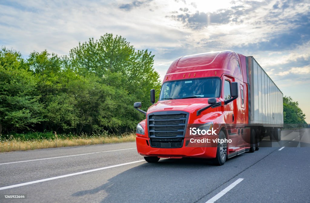 Red big rig long haul semi truck with black grille transporting cargo in dry van semi trailer running on the wide highway road Heavy loaded classic red big rig semi truck with high roof transporting commercial cargo at dry van semi trailer running on the straight wide divided multiline highway road for timely delivery Truck Stock Photo