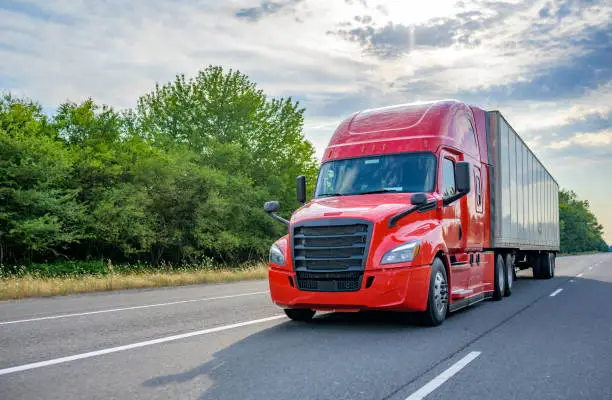 Photo of Red big rig long haul semi truck with black grille transporting cargo in dry van semi trailer running on the wide highway road