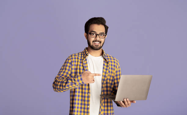 istock Online work, education, communication. Happy Indian guy pointing at laptop over lilac background 1265924451