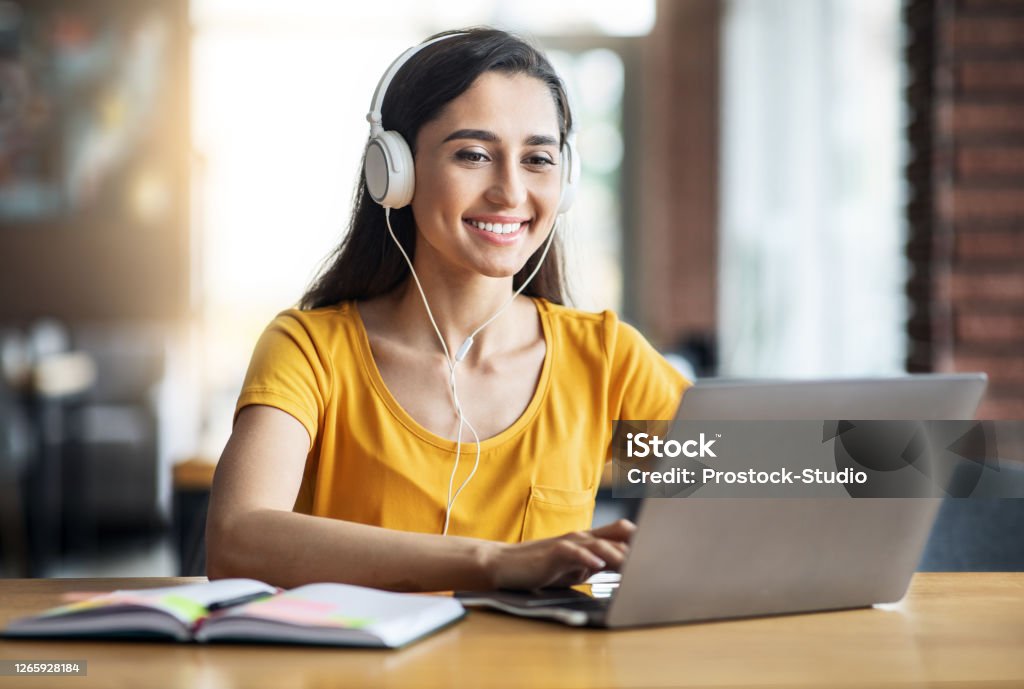 Smiling arab girl with headset studying online, using laptop Smiling arab girl with headset studying online, using laptop at cafe, taking notes, copy space Learning Stock Photo
