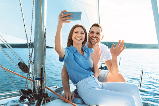 Joyful Spouses On Yacht Making Selfie On Phone And Waving Hand Sailing On Sea Outdoors. Yachting On Sailboat. Copy Space