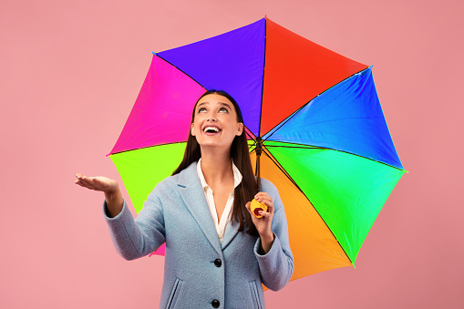 Portrait of excited beautiful woman standing under her umbrella and checking rain with open palm, looking up