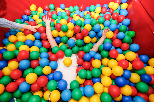 A child lying on back, arms outstretched, face covered with balls in a ball pool.