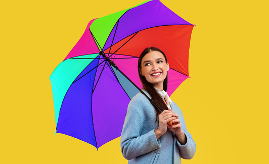 Playful Mood. Young carefree woman standing with rainbow umbrella and looking aside, yellow wall