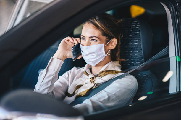uma jovem senta em um carro com uma máscara no rosto e fala em um celular, vida durante uma pandemia de vírus - driving mobile phone car talking - fotografias e filmes do acervo