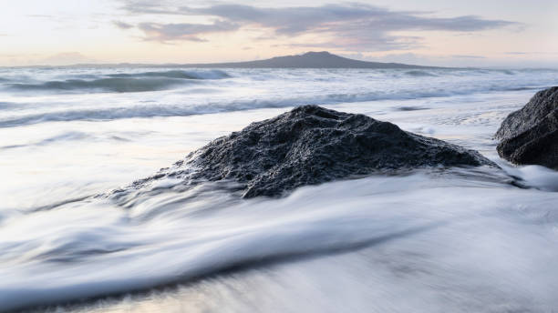 Waves crashing over rocks at sunrise with Rangitoto island in the distance. Image taken using slow shutter speed with the effect of silky water effect. Waves crashing over rocks at sunrise with Rangitoto island in the distance. Image taken using slow shutter speed with the effect of silky water effect. rangitoto island stock pictures, royalty-free photos & images