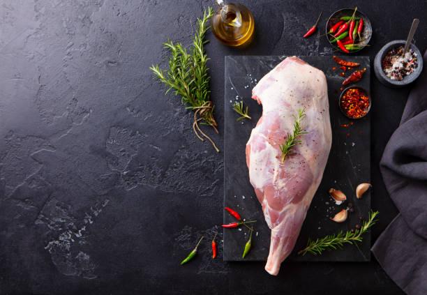 Raw lamb leg with spices and herbs on marble cutting board. Dark background. Copy space. Top view.