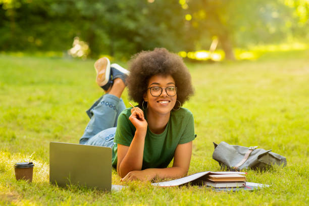estudante negra estudando ao ar livre com laptop e livros de trabalho, deitado no gramado - laptop computer grass nature - fotografias e filmes do acervo