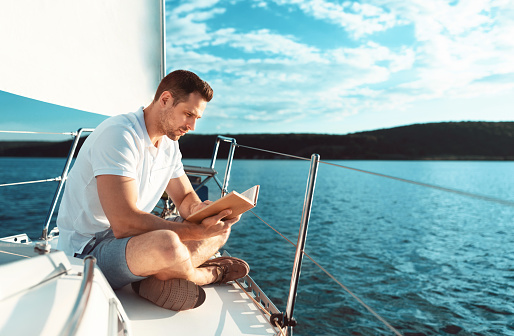 Dream Vacation On Yacht. Man Reading Book Relaxing Sitting On Sailboat Deck Sailing In Sea. Free Space For Text