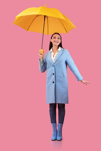 Enjoy The Weather. Cheerful stylish girl wearing autumn clothes srtanding with parasol over pastel pink studio wall