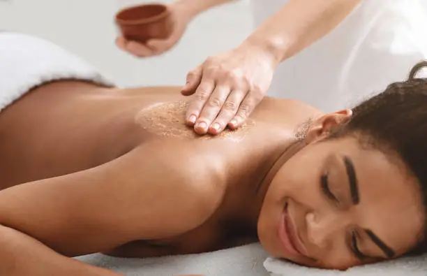 Beauty procedures at spa. Masseuse applying body scrub on black girl back, closeup