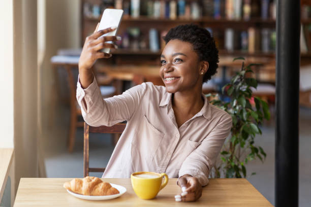 atractiva chica afroamericana tomando selfie con el teléfono móvil mientras desayuna en la cafetería - croissant telephone coffee shop on the phone fotografías e imágenes de stock