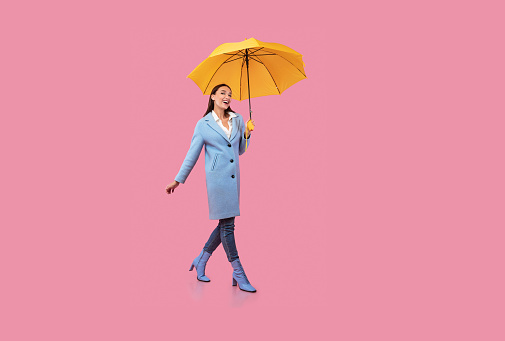Nice Day. Excited young model in blue coat walking with bright umbrella, isolated on pastel pink studio wall, copy space