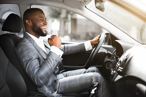 Young emotional black businessman in suit driving after successful business meeting, celebrating success, side view