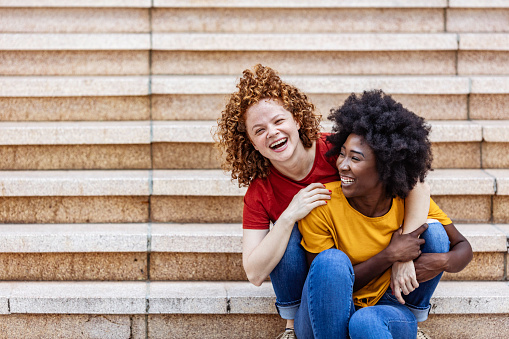 Two happy multi ethnic female friends Couple Hugging Each Other while sitting on steps outdoors .Two diverse Lesbian Women Outdoors. LGBT Concept.