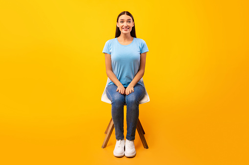 Studio shot of young woman sitting on the chair and looking at camera, copy space, orange wall