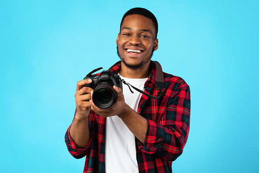 Photography. Black Photographer Guy Holding Modern Camera Posing On Blue Background. Studio Shot