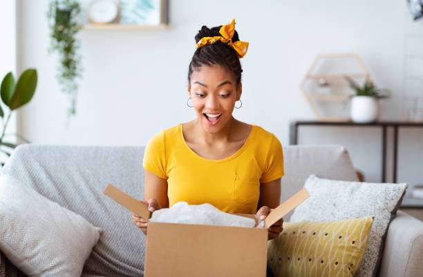 Happy girl unpacking clothes after online shopping Buying Via Internet. Excited afro girl sitting on sofa unboxing cardboard delivery package, copy space unpacking photos stock pictures, royalty-free photos & images