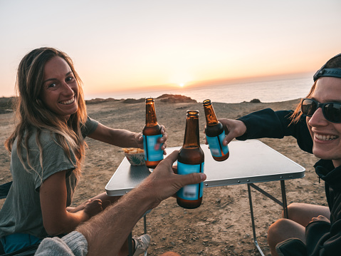 Three friends holding up beers cheering at sunset while sitting on camping chairs