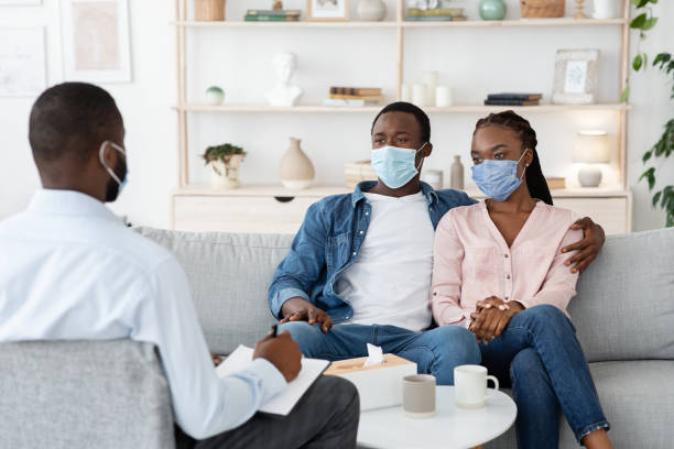 family therapist talking to black couple sitting in protective masks on couch at office - mental health professional family couple psychiatrist imagens e fotografias de stock