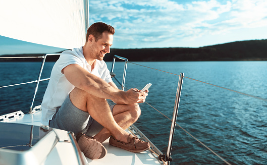 Yachting Concept. Man Using Smartphone App Sitting On Deck Relaxing Sailing Across The Sea Outdoors. Copy Space