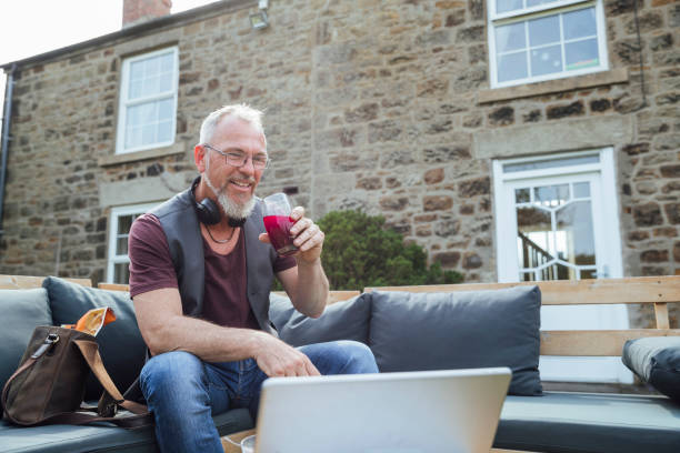 Taking a Break From Working A mature Caucasian man having a video call on a digital tablet while sitting outdoors on a bench with cushions. He is taking a break from working from home and drinking beetroot juice out of a glass. common beet audio stock pictures, royalty-free photos & images