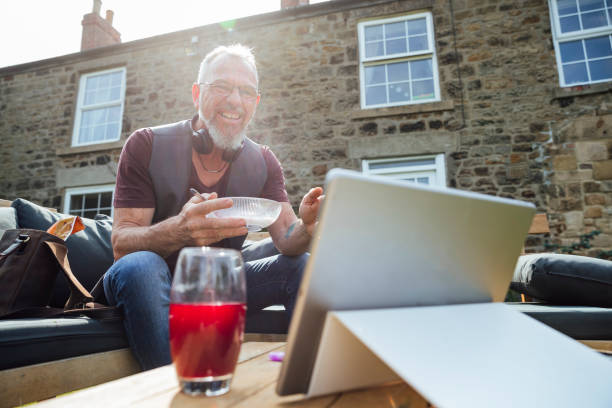 A Lovely Setting for Business A mature Caucasian man having a video call on a digital tablet while sitting outdoors on a bench. He is multi-tasking by working and eating at the same time. common beet audio stock pictures, royalty-free photos & images
