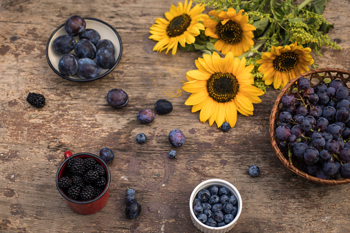 Directly above of wooden table with different purple fruits like grapes, plums, blueberries and blackberries. Sunflowers are placed to decorate a summer afternoon.