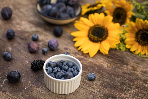 Close up of a small bowl of blueberries placed on a wooden table next to plums and sunflowers.